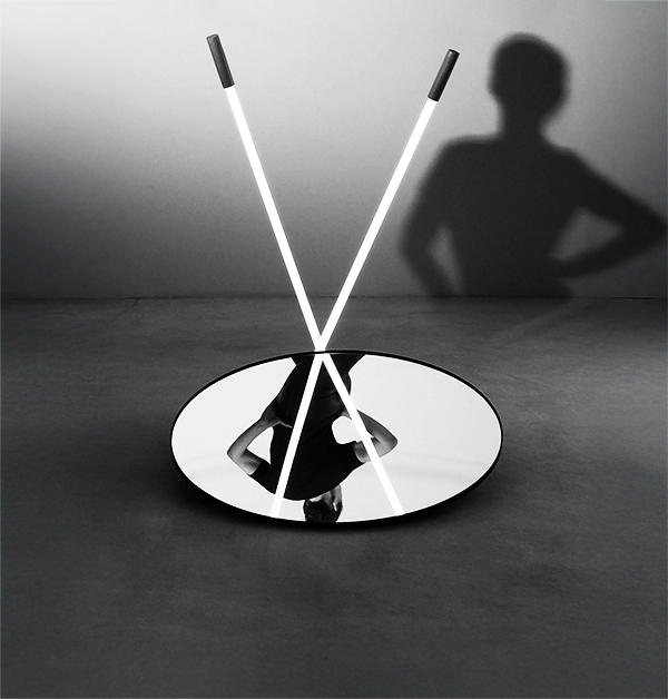 Ett runt spegelglas på golvet reflekterar en person som står bredvid. Två lysrör står lutade mot spegeln och bildar ett "V", vilket skapar en skugga av personen på väggen.