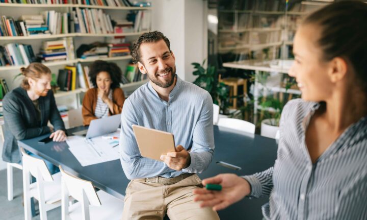 Fyra personer i ett modernt kontorsutrymme, läser och diskuterar vid ett bord med böcker i bakgrunden.