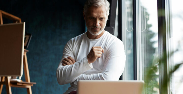 A man with grey hair and a beard, wearing a white shirt, stands with crossed arms, looking thoughtfully at a computer screen. He is near a window with natural light.