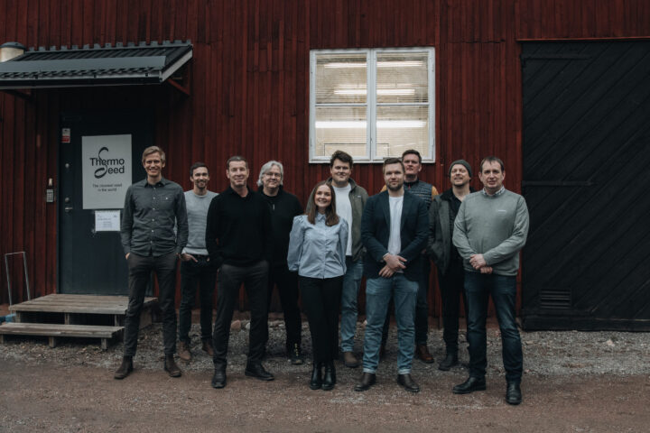 En grupp människor står framför en röd byggnad med en skylt som säger "Thermo Seed". Gruppen består av nio personer, som alla är klädda i avslappnade kläder.