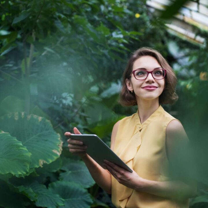 A woman with short hair and glasses is holding a tablet, standing amidst lush green plants, wearing a yellow top and looking upwards with a thoughtful expression.