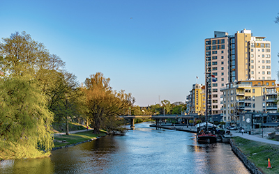 Stadsvy med en flodkant, grönskande träd och moderna flerbostadshus vid vattnet under en klarblå himmel.