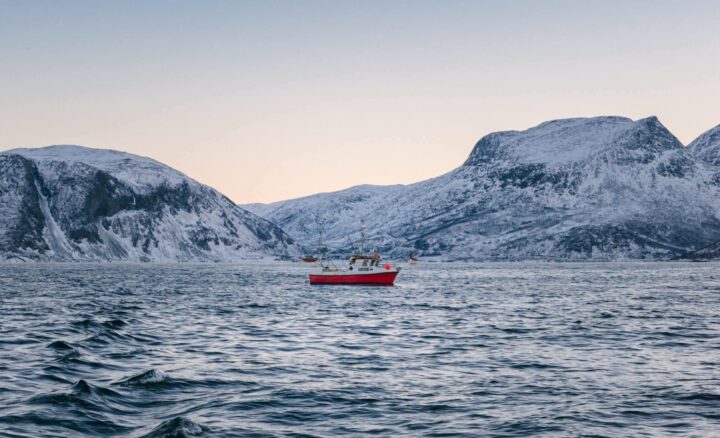 A red fishing boat on a calm, icy sea with snow-covered mountains in the background under a clear sky.