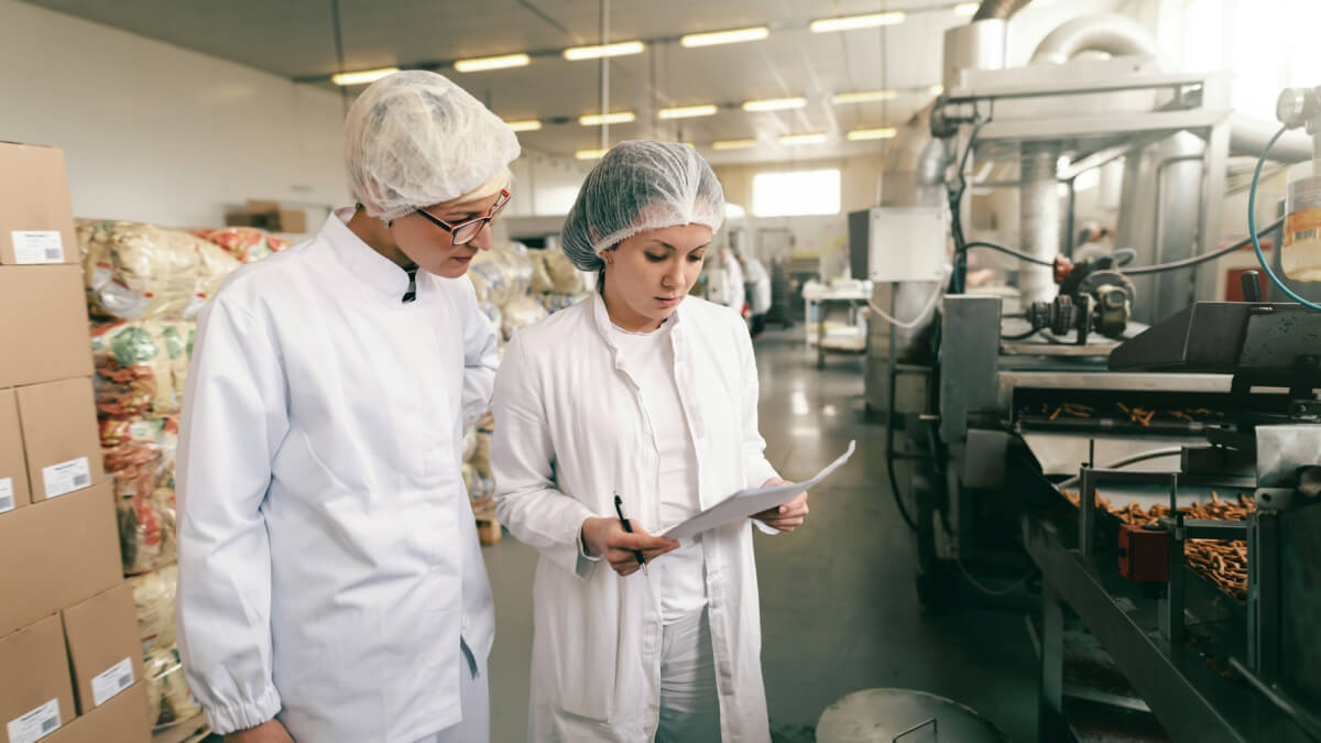 Two people in white lab coats and hairnets are reviewing a document in a food processing facility. They stand near machinery with products visible on the conveyor belt.
