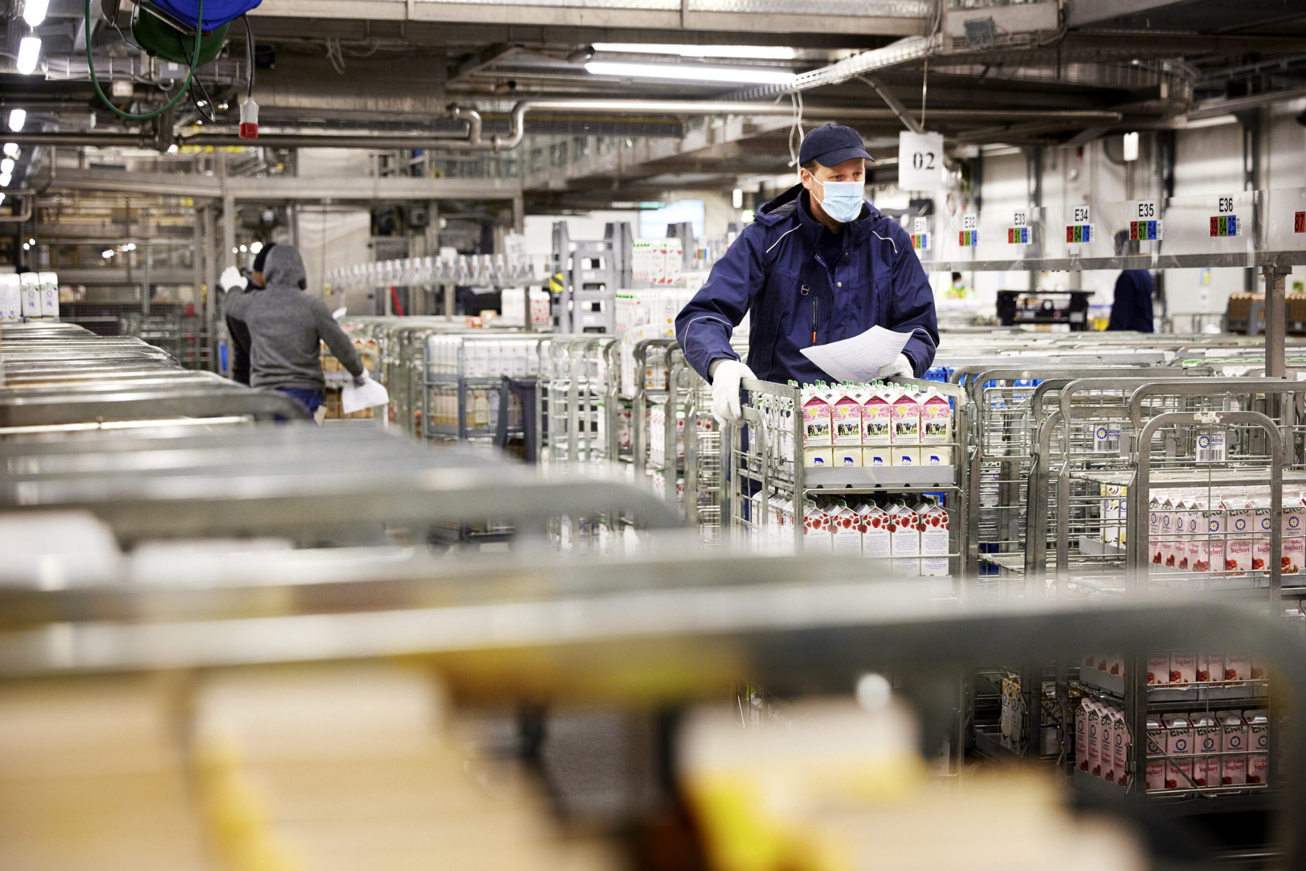 Worker in a warehouse wearing a mask and gloves, organising products in a trolley, with shelves in the background.