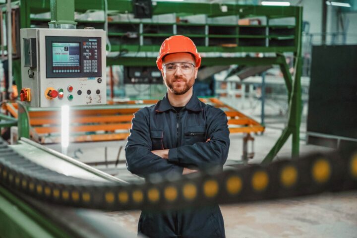 Man i orange hjälm och arbetsdräkt står framför maskin i fabrik.