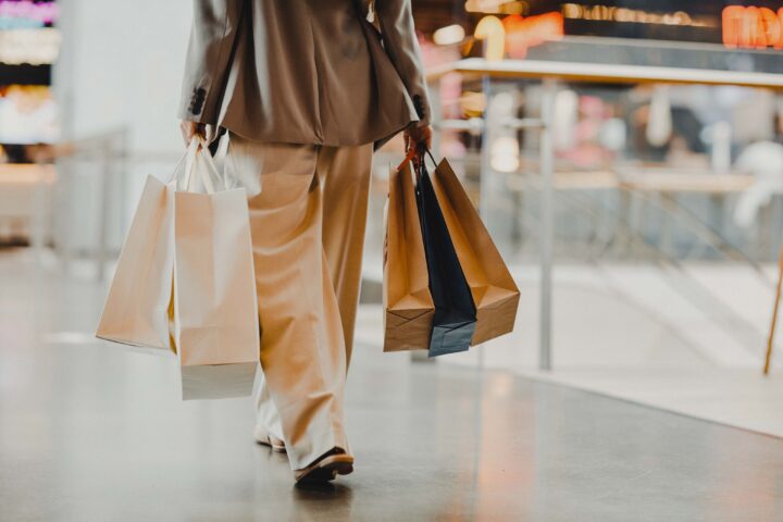 Person walking with shopping bags in a mall, wearing beige trousers and a jacket.