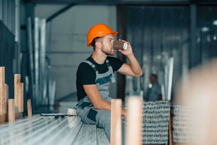 En byggnadsarbetare med orange hjälm dricker kaffe på sittande.