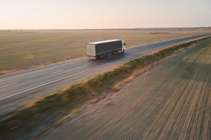 A lorry drives down a long, empty highway through open fields at sunset.