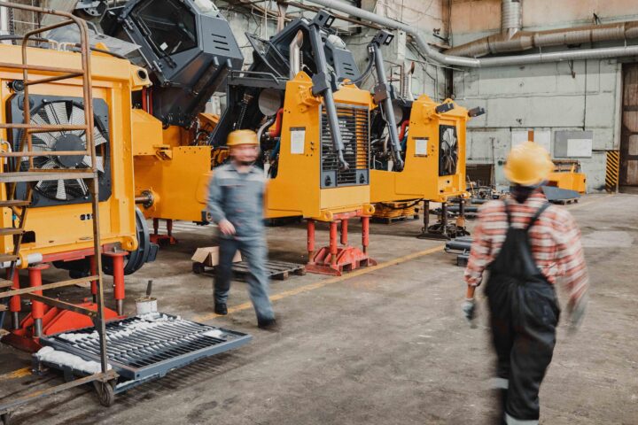 Arbetare i skyddshjälmar rör sig genom en industrilokal med stora gula maskiner.