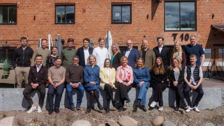 En grupp människor poserar utomhus framför en röd tegelvägg, några sitter och andra står. Soligt väder.