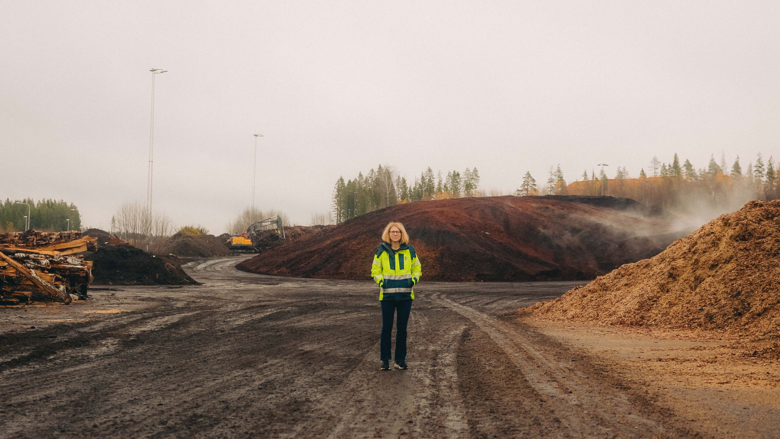 Person i gul jacka står i ett industriområde med jordhögar och träd i bakgrunden.