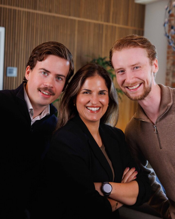 Tre personer ler framför en trävägg.