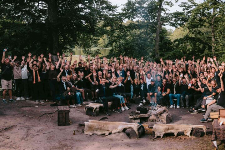 En stor grupp människor med händerna uppsträckta, utomhus i en skogsmiljö.