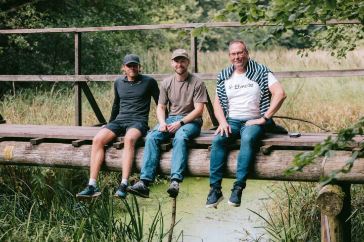 Tre personer sitter på en träbro över en grönskande åker.