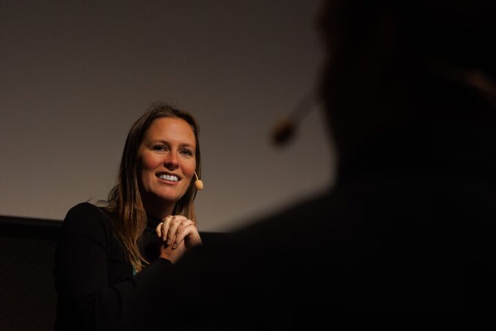 Kvinna med mikrofon talar på ett evenemang, mörk bakgrund.