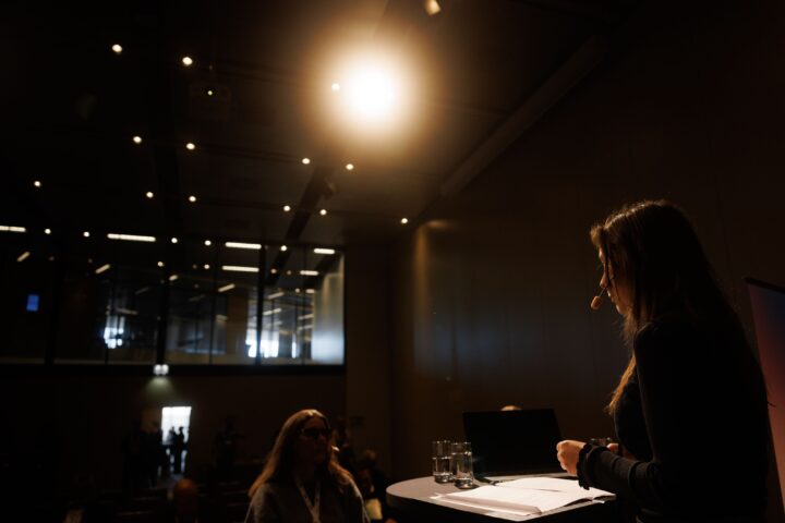 Kvinnlig talare vid ett podium i ett mörkt rum med publik.