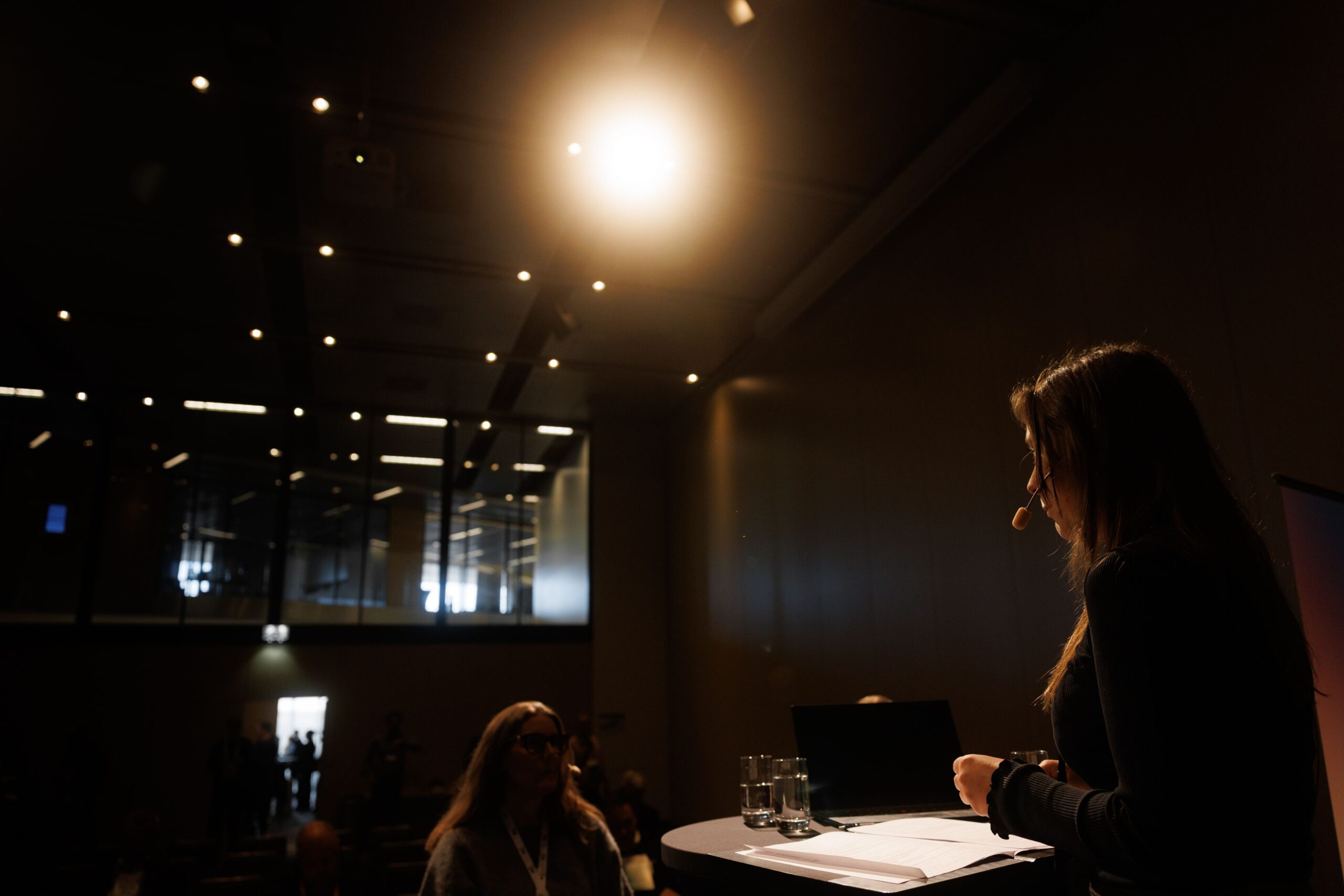 A person speaking with a headset microphone at a podium, in a dark room with spotlights on the ceiling.