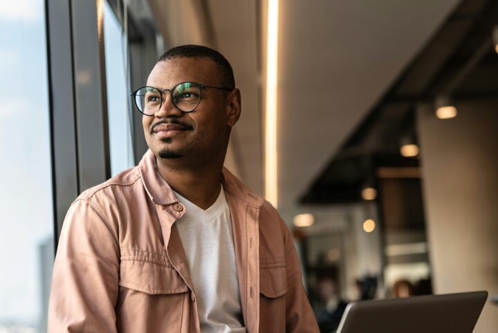 Man med glasögon i rosa jacka tittar ut genom fönster, suddig kontorsmiljö i bakgrunden.