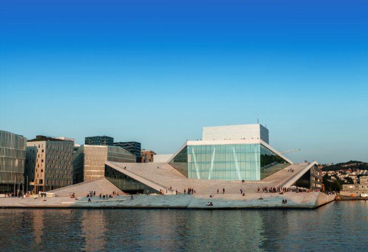 Modern waterfront building with angled glass facade, people walking and sitting around, clear blue sky.