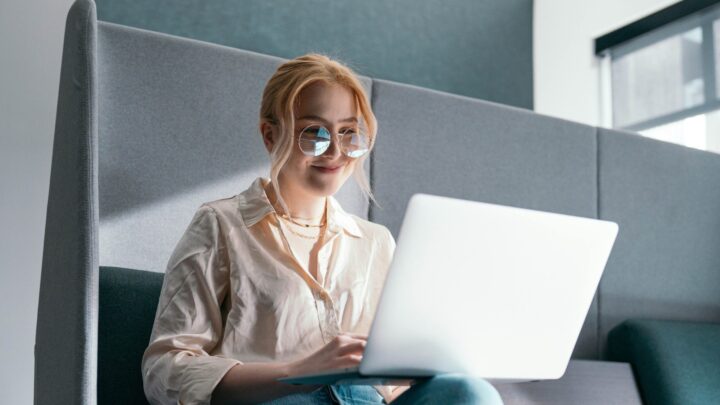 Woman with glasses using a laptop, sitting on a couch in a bright room.