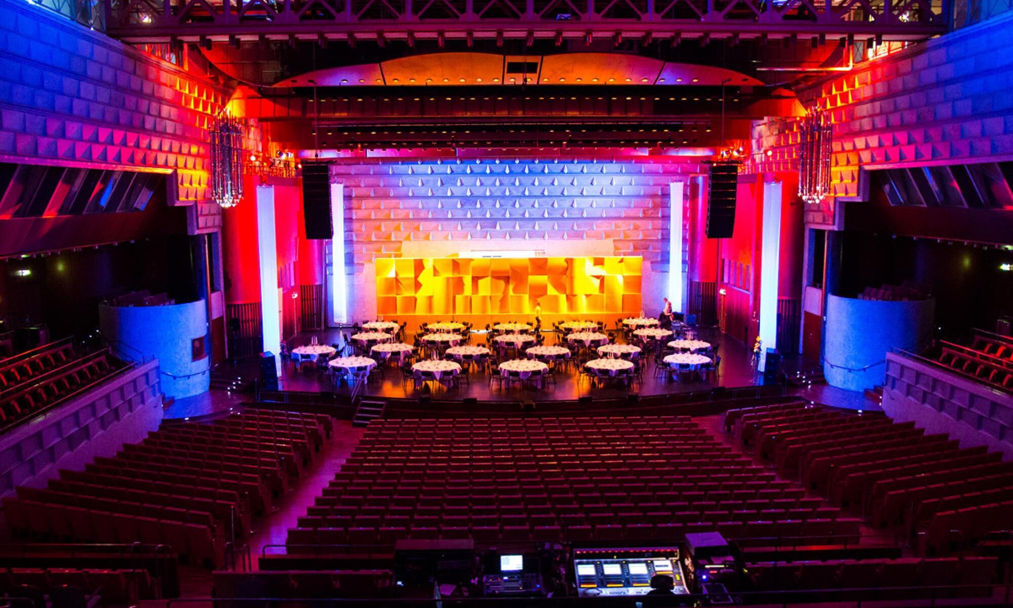 Färgglad inomhussal med runda bord uppställda på scenen och tomma röda stolsrader i förgrunden.