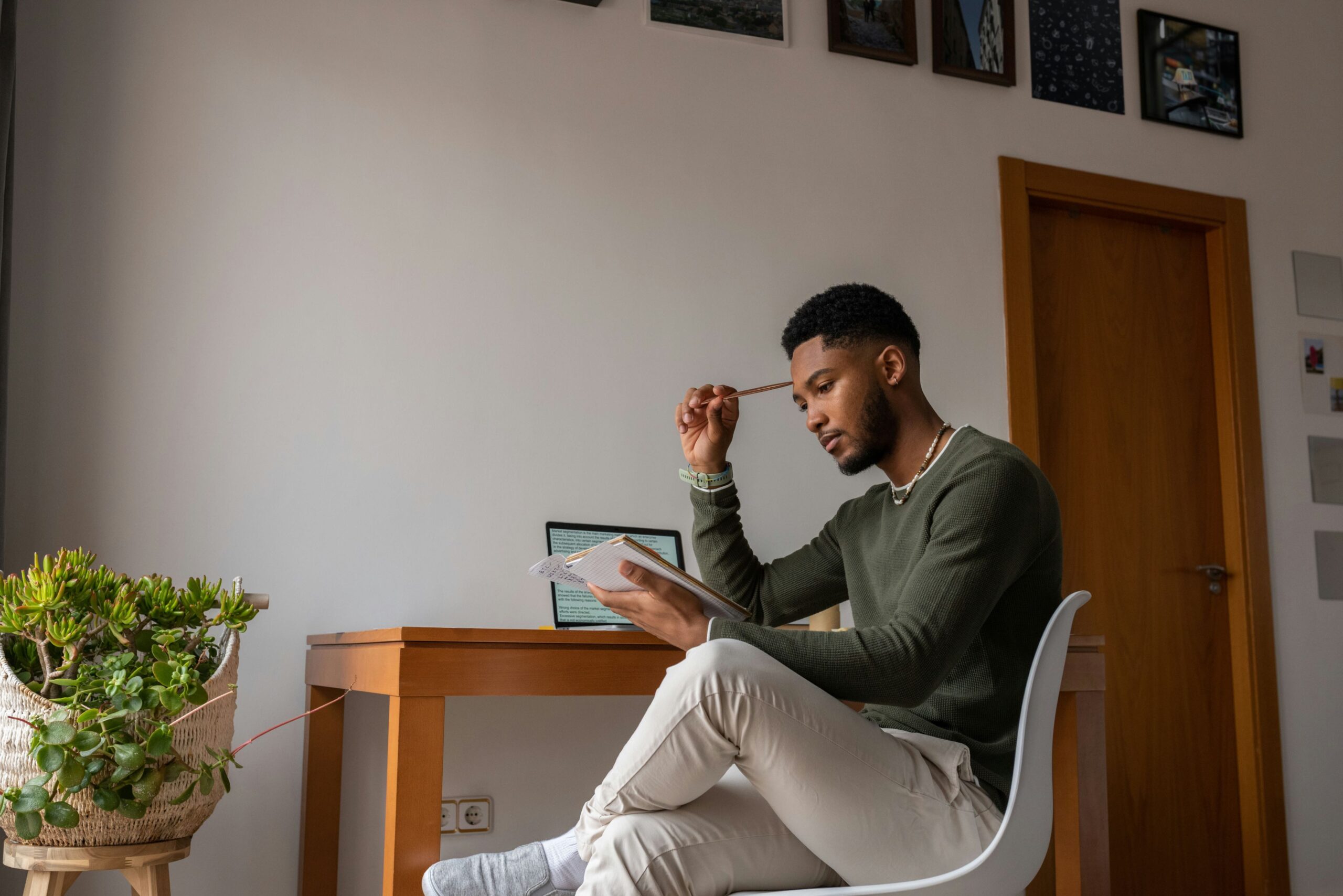 En person som skriver anteckningar vid ett bord, med en dator och en växt i bakgrunden.