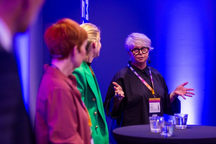 Three women in discussion on stage at a conference, one speaking, wearing glasses, with blue and purple lighting.