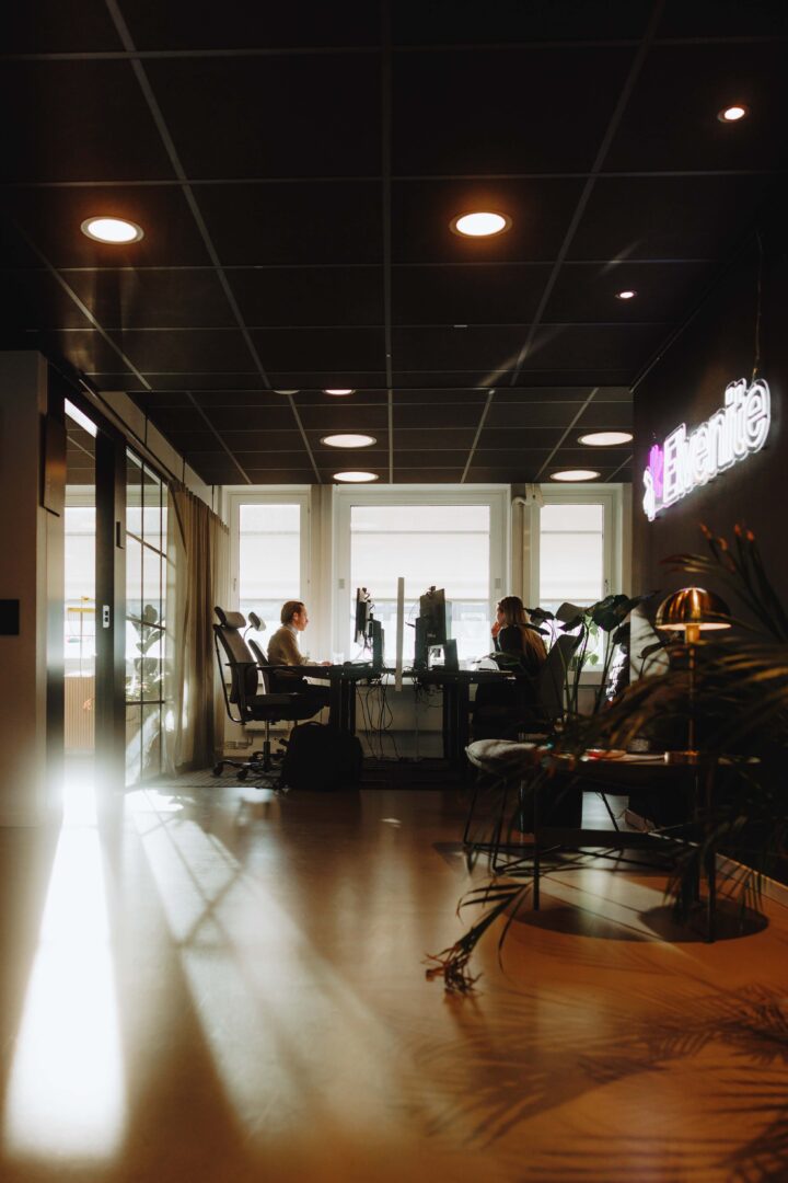 Modern office space with two people working at desks, large windows, plants, and a neon sign on the wall.