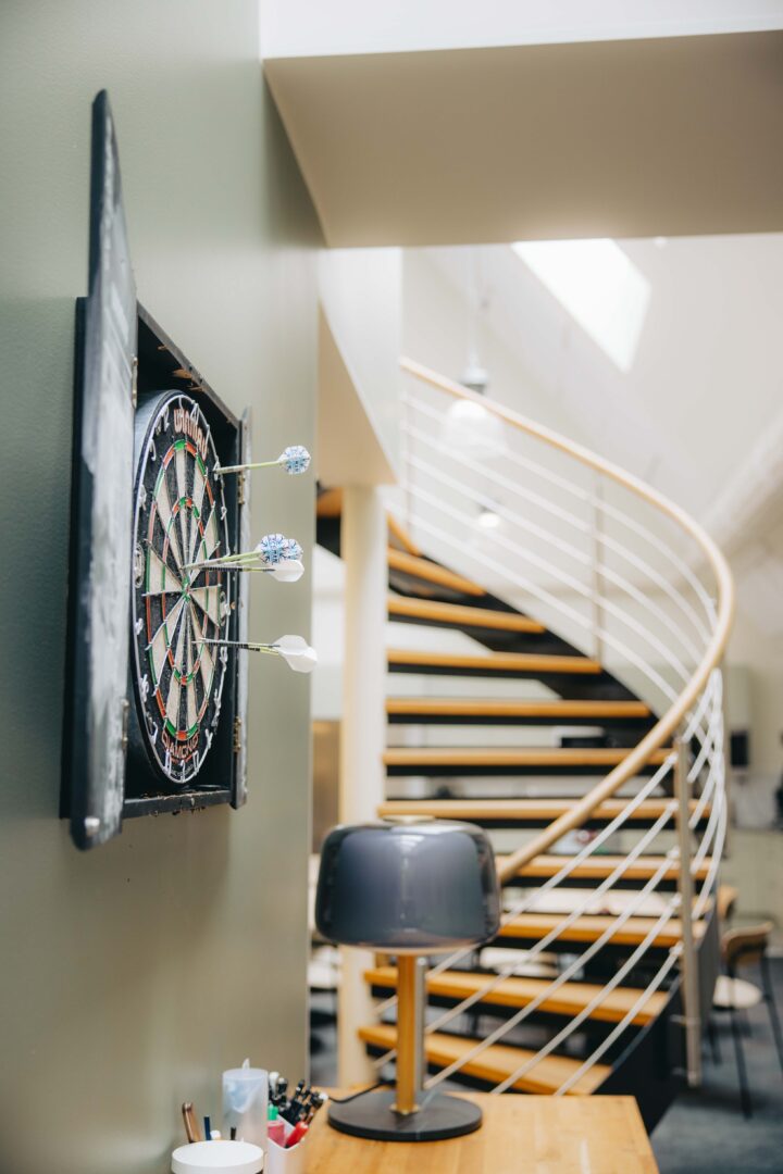 Dartboard with darts on a green wall, desk with lamp, and a spiral staircase in the background.