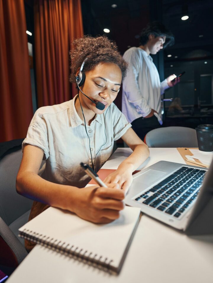 Woman wearing headset writes in notebook at laptop, man stands in background using phone in office setting.