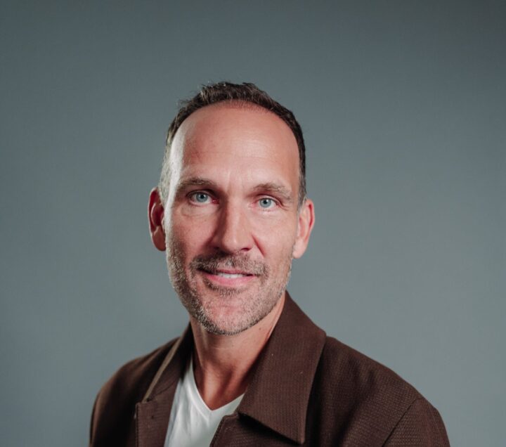 Man with short hair and stubble wearing a brown jacket and white shirt, posing against a plain grey background.