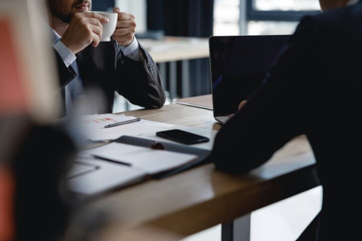 Two people in business attire sitting at a desk with papers, a laptop, and one holding a coffee cup.