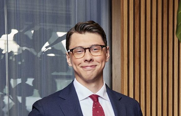 Ung man i kostym, vit skjorta, röd slips och glasögon står framför trälister och ett fönster, och ler.